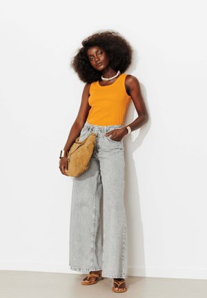 Femme aux cheveux afro naturels portant un haut orange sans manches, un jean large gris clair, des sandales marron clair et tenant un sac à main marron clair.
