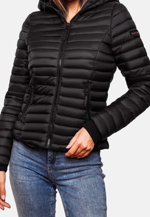 Frau trägt eine taillierte schwarze gesteppte Jacke mit durchgehendem Reißverschluss und Reißverschlusstaschen, kombiniert mit blauen Jeans.