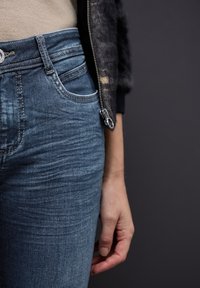 Jeans de mezclilla azul con un ajuste clásico, que presentan un desvanecimiento sutil y un bolsillo delantero. Mano descansando sobre los jeans, contra un fondo oscuro.