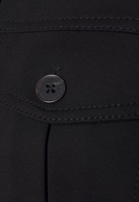 Mustaa kangasta, jossa kiiltävä napin yksityiskohta, olkaen ommeltu sauma ja pleatattu muotoilu. Sileä tekstuuri, lähikuva vaatekappaleen yksityiskohdista.