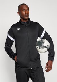 Jachetă sport neagră cu fermoar complet, accente albe pe mâneci, manșete cu nervuri și detaliu de logo. Modelul ține o minge de fotbal.