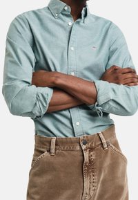 Homme à la peau foncée portant une chemise bleu clair boutonnée avec les manches retroussées et un pantalon marron, les bras croisés sur la poitrine.