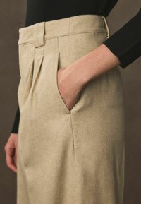 Beige linnedbukser med folder og sidelommer, der har en struktureret finish. Taljen er formet, og en sort sweater bæres ovenfor.