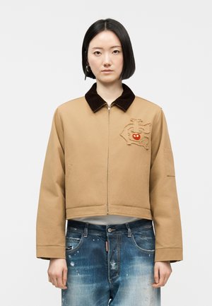 Fiatal nő, rövid fekete hajjal, bézs színű, cipzáras kabátot visel sötét gallérral és hímzett piros orr részlettel, amelyet kopott kék farmerral párosít.