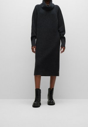 Person, die ein dunkelgraues, langärmliges Strick-Midikleid und schwarze Schnür-Ankle Boots trägt, steht vor einem schlichten hellen Hintergrund.