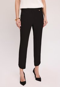 Czarne dopasowane spodnie o gładkiej teksturze, długości do kostek, z delikatnym plisowaniem z przodu. Posiadają mały złoty akcent w talii.
