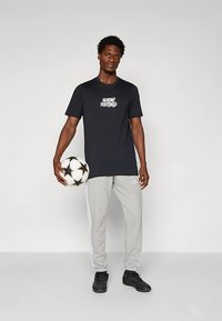 Schwarzes Baumwoll-T-Shirt mit "Adidas Football"-Logo, kombiniert mit grauer Jogginghose mit weißen Streifen. Modell hält einen schwarz-weißen Fußball.