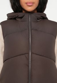 Gilet imbottito marrone con colletto alto, chiusura a zip e una texture liscia. Presenta cuciture sottili e un design leggero.