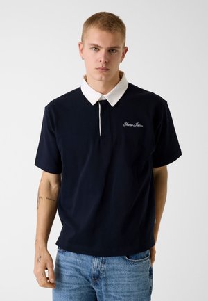 Giovane uomo con capelli biondi corti che indossa una polo nera con colletto bianco e logo "Guess Jeans", abbinata a jeans di denim blu.