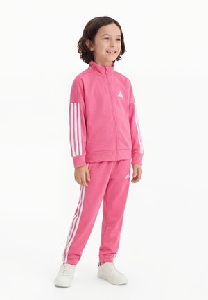Niño sonriente con chándal rosa de Adidas con rayas blancas, de pie y con zapatillas blancas sobre fondo blanco.