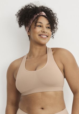 Lächende Frau mit lockigem Haar, rosa Stirnband und passendem Sport-BH, steht vor einer weißen Wand und schaut nach rechts.