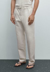 Pantaloni leggeri beige con vita elasticizzata e cordino, tasche laterali e vestibilità rilassata, abbinati a ciabatte nere.