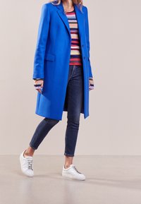 Blauer Wollmantel, doppelt knöpfbar mit Revers, über einem gestreiften Strickpullover getragen. Kombiniert mit dunklen Slim Jeans und weißen Sneakers.