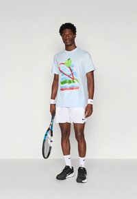 Světle modré tričko s krátkými rukávy a barevnou tenisovou grafikou, bílé šortky, černé tenisky a tenisová raketa. Hladká látka, volný střih.