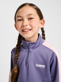 Jong meisje met gevlochten haar glimlachend, draagt een paarse Hummel Performance sportjas met ritssluiting tegen een effen lichte achtergrond.