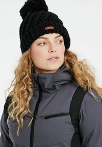Graue, gepolsterte Jacke mit schwarzen Akzenten und einer Reißverschlusstasche, kombiniert mit einer schwarzen Strickmütze mit Bommel und einem Logotag.