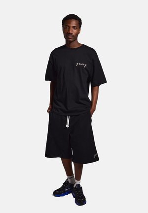 Camiseta de algodón negra con logotipo bordado, combinada con pantalones cortos negros con cordón ajustable. Zapatillas deportivas negras y azules.