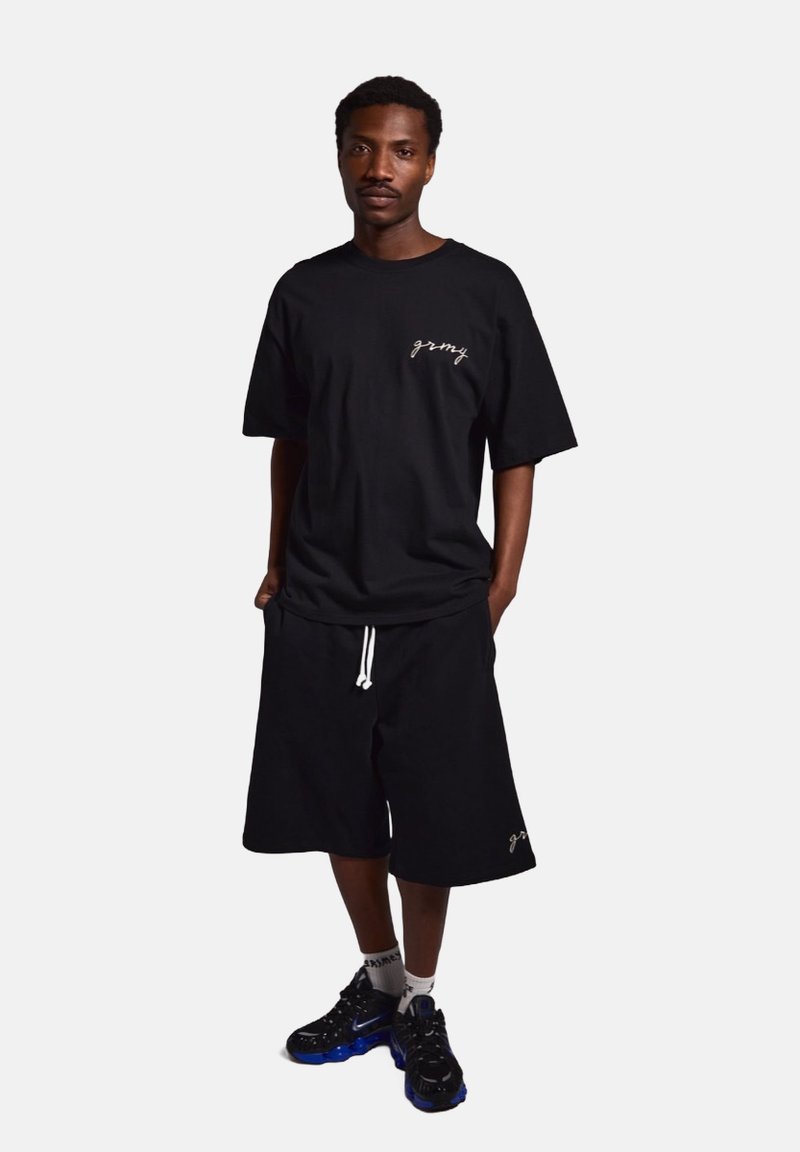 Camiseta de algodón negra con logotipo bordado, combinada con pantalones cortos negros con cordón ajustable. Zapatillas deportivas negras y azules.
