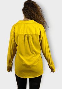 Camisa amarilla de botones con mangas largas, detalle de pliegue en la parte trasera y diseño recto. Textura suave y acabado de tela blanda.
