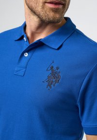 Muž v modrém polo tričku s vyšitým logem dvou jezdců na koních na hrudi, detailní pohled.