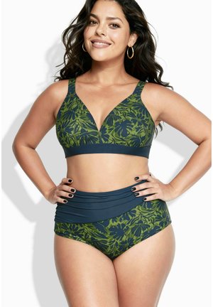 Femme aux cheveux bouclés et foncés portant un bikini floral vert et marine avec un bas taille haute, debout les mains sur les hanches, souriant devant un fond blanc.