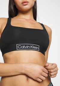Zwarte katoenen sportbeha met een vierkante halslijn en een brede elastische band met het witte "Calvin Klein" logo. Gladde textuur, eenvoudig ontwerp.