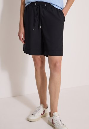 Person iført sorte shorts med snøre og hvide sneakers med lilla detaljer står mod en ensfarvet lys baggrund.