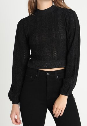 Femme portant un crop top noir texturé à manches longues avec des épaules bouffantes et un pantalon noir taille haute sur un fond uni.
