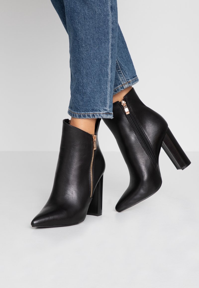 Bottes noires en cuir à talons hauts avec fermetures éclair latérales portées avec un jean en denim bleu sur une surface blanche.
