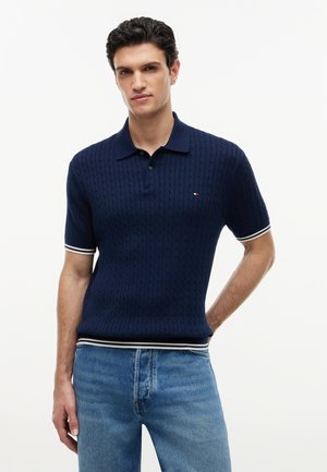 Mand iført en marineblå kortærmet strikket polo med hvide stribede kanter og lyseblå jeans mod en hvid baggrund.