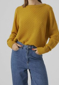 Senfgelber gestrickter Pullover mit Rautenmuster, rundem Ausschnitt und gerippten Bündchen, kombiniert mit hoch taillierter blauer Jeans.