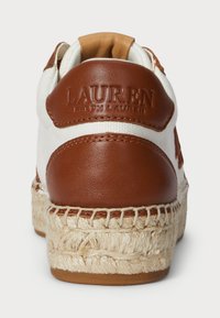 Biele plátené tenisky s hnedou koženou pätou a logom, so splietanou jutovou medzi podrážkou a štruktúrovanou gumovou podrážkou.