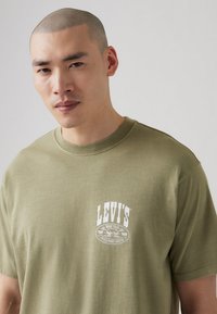 Olive green cotton t-shirt with a round neckline and short sleeves, featuring a white printed logo on the chest. Smooth texture.
