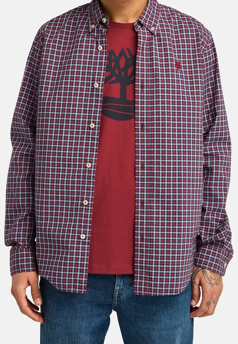 Homme portant une chemise à carreaux bleue, rouge et blanche boutonnée sur un t-shirt rouge avec un logo d'arbre noir, associée à un jean bleu.