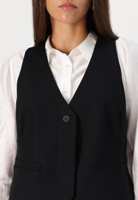 Black vest with a smooth texture, featuring a V-neck, single button closure, and two front pockets, layered over a white shirt.