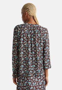 Blusa marrone scuro a maniche lunghe con un motivo floreale in blu e crema, caratterizzata da un colletto arricciato e una morbida texture del tessuto.