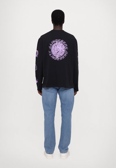 Schwarzes Langarmshirt mit lila grafischem Design auf dem Rücken, rundem Logo und weiteren Grafiken auf den Ärmeln. Kombiniert mit hellblauen Jeans.