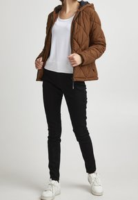 Veste matelassée brune avec capuche, portée par-dessus un haut blanc, associée à un pantalon noir ajusté et des baskets blanches. Texture lisse, design sportif.