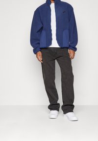 Blå fleecjacka med kontrasterande blå detaljer och fickor, bärs över en vit t-shirt, tillsammans med mörkgrå byxor och vita sneakers.