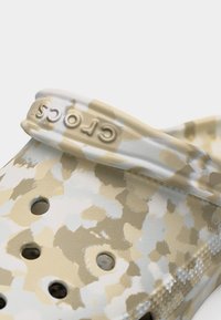 Κοντινή λήψη ενός Crocs με μοτίβο παραλλαγής σε μπεζ και άσπρο, με τρύπες αερισμού και το λογότυπο Crocs στο λουράκι.