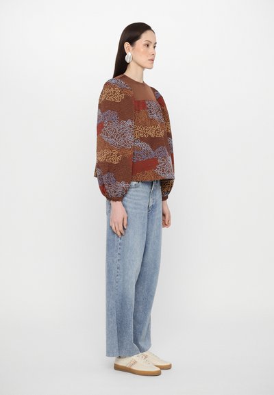 Brun mönstrad blus med puffärmar, ljusblå jeans och beige sneakers med flerfärgade ränder. Byxorna har rak skärning.