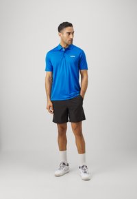 Blå polo med korte ærmer, prydet med et lille hvidt logo, parret med sorte shorts og hvide sneakers med grå detaljer.