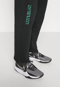 Schwarze Jogginghosen mit dem in Türkis gestickten Schriftzug "REWARD" am Bein, kombiniert mit schwarz-grauen Sportschuhen, die ein strukturiertes Design und weiße Akzente aufweisen.