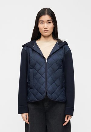 Mladá žena v tmavomodrej prešívanej bunde na zips s kapucňou a dlhými rukávmi, oblečená cez béžové tričko, stojaca pred jednoduchým svetlým pozadím.