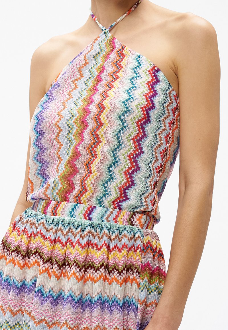 Kvinde iført en flerfarvet halterneck-top med zigzag-mønster og matchende højtaljet nederdel i klare, varierede farver og tekstureret stof.