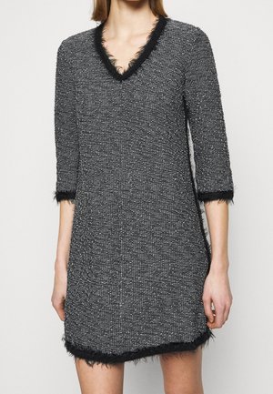 Kobieta w czarno-białej, teksturowanej sukience z dekoltem w kształcie litery V, z rękawami 3/4 i piórkowymi zdobieniami na dekolcie, mankietach i dole sukienki.