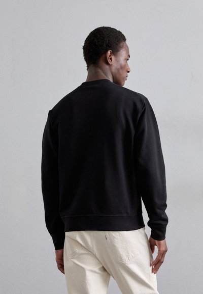 Black sweatshirt with a crew neckline and ribbed cuffs, designed with a relaxed fit. Worn with light-colored pants, showing a simple, clean look.