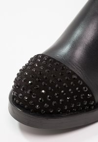 Chaussure noire avec un tige en cuir lisse et un capuchon en suede noir texturé orné de clous noirs brillants, présentant un bout rond.