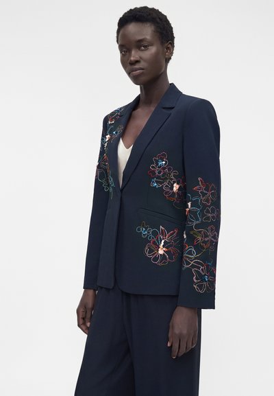 Μαύρη γυναίκα φοράει σκούρο μπλε σακάκι με πολύχρωμη κεντητή λουλουδάτη διακόσμηση στα μανίκια και μπροστά, σε συνδυασμό με αντίστοιχο μπλε παντελόνι και λευκό τοπ.