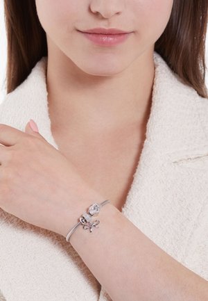 Silver bracelet with a smooth, flexible band and two decorative charms—one spherical and one bow-shaped, featuring intricate details.